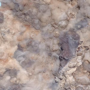 May include: Close-up of a natural stone with a textured surface. The stone features a mix of colors, including beige, gray, and hints of purple. The surface has a bubbly, uneven texture.