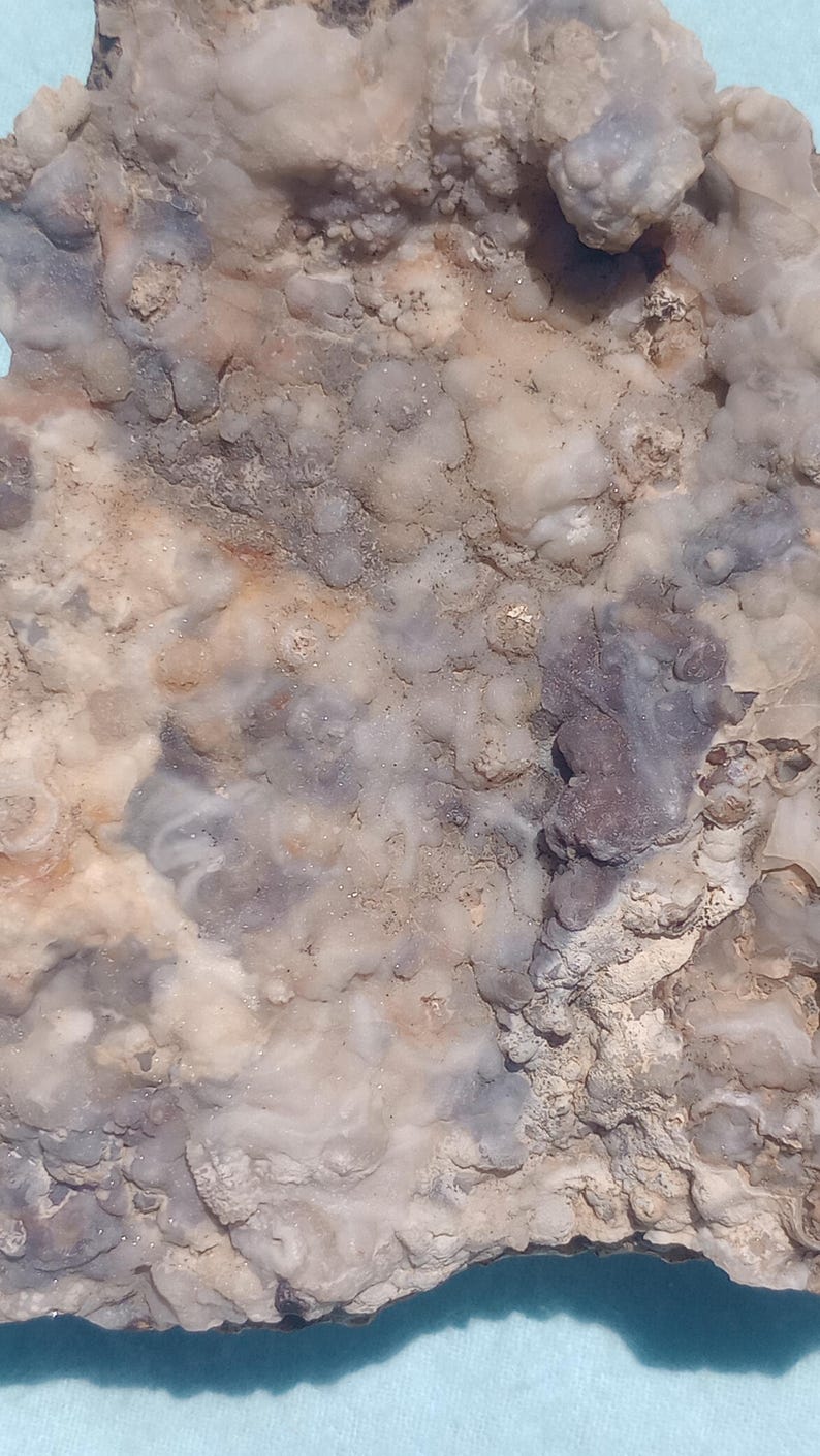 May include: Close-up of a natural stone with a textured surface. The stone features a mix of colors, including beige, gray, and hints of purple. The surface has a bubbly, uneven texture.
