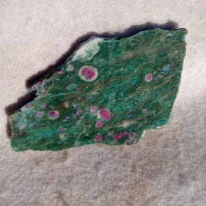 May include: A rough-cut gemstone with a vibrant green base color, speckled with numerous small, round, deep pink and red inclusions. The stone has a natural, irregular shape and a slightly polished surface, showcasing its unique mineral composition.