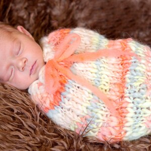 May include: A newborn baby sleeps peacefully, swaddled in a colorful, hand-knitted blanket. The blanket features pastel shades of pink, yellow, and blue, tied with a matching ribbon. The baby rests on a soft, brown, faux fur surface.