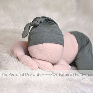Puede incluir: Un gorro de punto gris con un nudo en la parte superior, usado por un muñeco de bebé de peluche acostado sobre una superficie blanca y esponjosa. El muñeco lleva una camisa gris de manga larga.