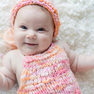 Puede incluir: Un bebé con un gorro de punto rosa y naranja y un top de punto sin mangas a juego. El bebé está tumbado sobre una manta blanca y esponjosa.