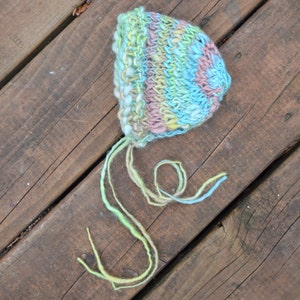 Pode incluir: Um chapéu de bebé de crochê em tons de azul, verde e rosa com uma longa gravata atada.