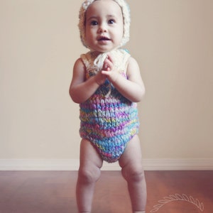 May include: A baby wearing a colourful crocheted onesie and a white crocheted hat. The baby is standing on a wooden floor and looking up.
