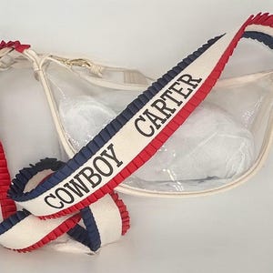 May include: A clear, cream-colored crossbody bag with a strap featuring red, white, and blue pleated trim. The strap has the text "COWBOY CARTER" in black. The bag has a zipper closure and contains a white object.