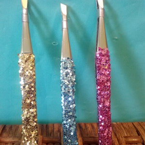 May include: Three pairs of tweezers with glitter handles. The tweezers are silver with a slanted tip. The handles are gold, blue, and pink.