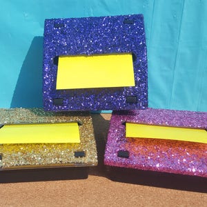 May include: Three glitter-covered note dispensers with yellow sticky notes. The dispensers are purple, gold, and pink.
