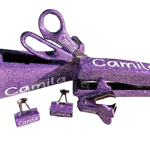 May include: A set of purple glitter office supplies, including scissors, a stapler, and binder clips. The name "Camila" is printed in white on each item. The items are set against a white background.