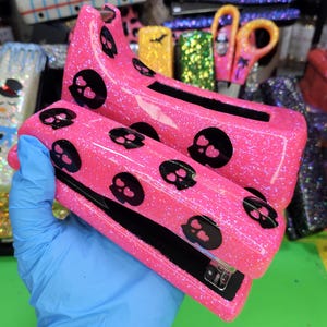 May include: A pink glitter stapler with black skull designs. The stapler is held by a gloved hand, showcasing its sparkly texture and decorative skulls. Other decorative office supplies are visible in the background.