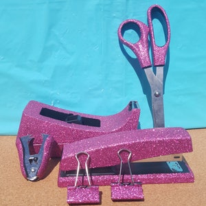 May include: A set of pink glitter office supplies including a stapler, a tape dispenser, a pair of scissors, and two binder clips.