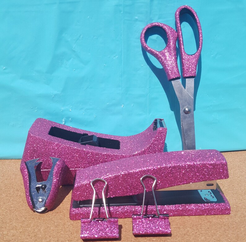 May include: A set of pink glitter office supplies including a stapler, a tape dispenser, a pair of scissors, and two binder clips.