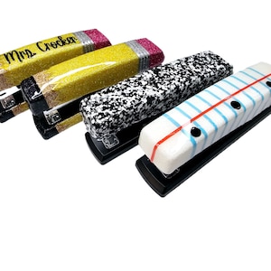 May include: Four staplers decorated with different school-themed designs. The staplers are black with a silver staple remover. The designs include a yellow pencil with glitter, a black and white speckled design, a pink and white striped design, and a blue and white lined paper design.