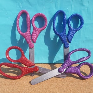 May include: Three pairs of glitter scissors in pink, blue, and purple. The scissors are all closed and have a silver handle.