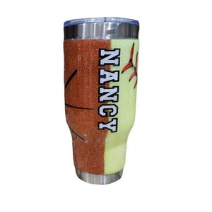 May include: A stainless steel tumbler with a glitter finish. One side is orange with a baseball design and the other side is light green with the name "NANCY" in white letters.