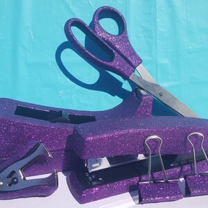 May include: Purple glitter desk organizer set with a stapler, staple remover, scissors, and two binder clips.