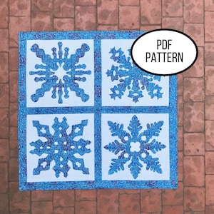 May include: A blue and white quilt block with four snowflake designs. The quilt block is on a red brick surface. The text "PDF PATTERN" is in a white circle.