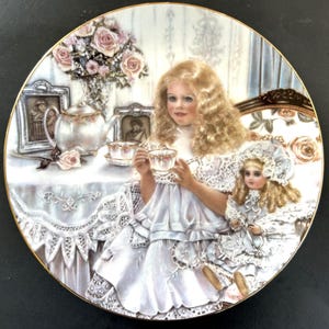 May include: A porcelain plate with a gold rim features a painting of a young girl in a white dress holding a teacup. She is sitting at a table with a teapot and a doll. The background includes a vase of pink roses and a window with a blue curtain.