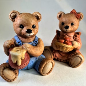May include: Two ceramic bear figurines. The left bear is light brown, wearing blue overalls and holding a red pot with yellow honey dripping down the side, with the word HONEY embossed on it. The right bear is light brown, wearing a peach dress and a red bow, holding a basket of red apples.