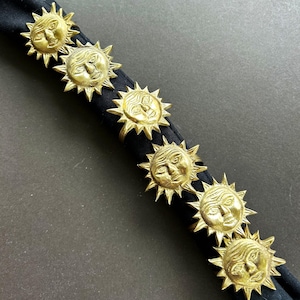 May include: Six gold-colored sun-shaped napkin rings. Each ring features a detailed sun face with radiating spikes. The rings are arranged on a black napkin, creating a striking contrast. These decorative rings add a touch of elegance to any table setting.