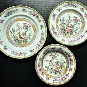 May include: Three white porcelain plates with a green and gold trim. The plates feature a floral pattern with a tree branch and pink flowers.