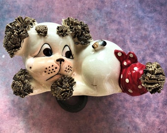 May include: A ceramic dog figurine in a reclining pose. The dog is primarily white with brown accents on its ears and head. It has a red bow with white polka dots and a black wheel. The dog's face has black eyes and a black nose.