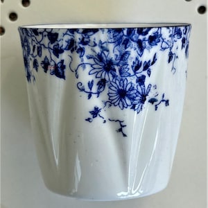 May include: White ceramic cup with a blue floral design. The cup features a repeating pattern of blue flowers and vines around the rim. The cup has a slightly curved shape and a smooth, glossy finish.