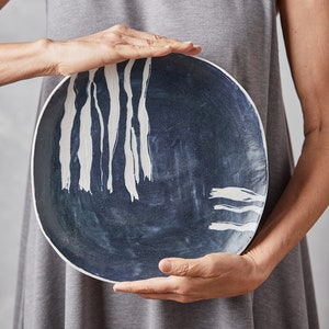 Puede incluir: Un plato de cerámica redondo con un centro azul oscuro y pinceladas verticales blancas abstractas. El plato tiene un borde blanco y lo sostiene una persona que lleva un vestido gris.