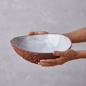 Op de afbeelding: Een witte keramische kom met een ruw, getextureerd exterieur. De kom wordt in twee handen gehouden.