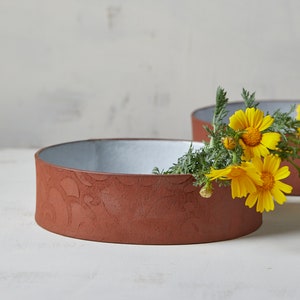 May include: Two low, round, terracotta-colored ceramic bowls with a textured surface. The interior of the bowls is a light gray. Yellow flowers and green foliage are arranged in one of the bowls. The bowls are on a white surface.