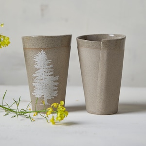 Puede incluir: Dos tazas de cerámica, una con un diseño de árbol blanco, ambas con un esmalte beige moteado.