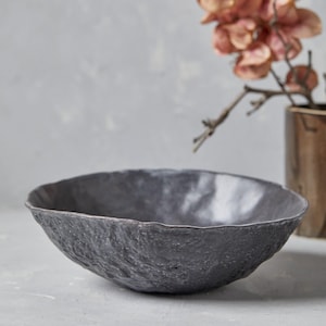 May include: A dark grey, textured bowl with an organic, irregular rim. The bowl has a rough, stone-like appearance. A bronze-coloured vase with dried flowers is in the background, adding a touch of natural decor.