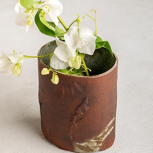 May include: A cylindrical, textured, brown ceramic vase with a green interior holds white bougainvillea flowers and green leaves. The vase has a rough, organic texture and a slightly irregular shape, suggesting a handmade quality.