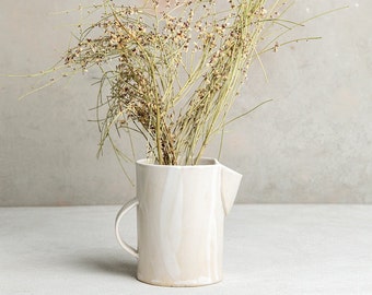 May include: A cream-colored ceramic pitcher with a handle and spout, holding dried, wispy, beige-colored stems with tiny brown buds. The pitcher has a marbled effect and sits on a light gray surface.
