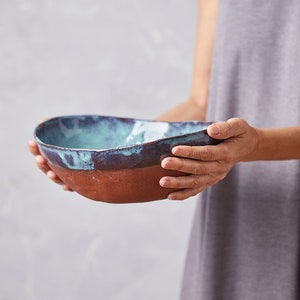 May include: A large, oval-shaped ceramic bowl with a teal glaze and a brown rim. The bowl is being held by a person's hands.
