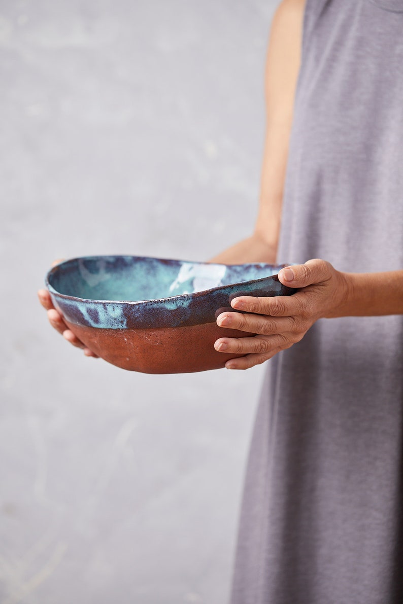 May include: A large, oval-shaped ceramic bowl with a teal glaze and a brown rim. The bowl is being held by a person's hands.