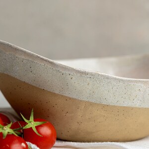May include: A large, off-white ceramic bowl with a brown rim. The bowl has a speckled glaze and is sitting on a white surface. There are red tomatoes in the foreground.