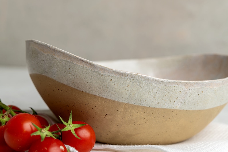 May include: A large, off-white ceramic bowl with a brown rim. The bowl has a speckled glaze and is sitting on a white surface. There are red tomatoes in the foreground.
