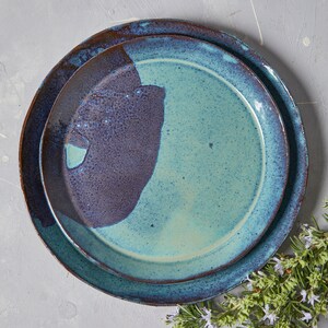 May include: Two ceramic plates, one nested inside the other. The top plate is a light blue with a dark blue and brown glaze. The bottom plate is a dark blue with a brown glaze.