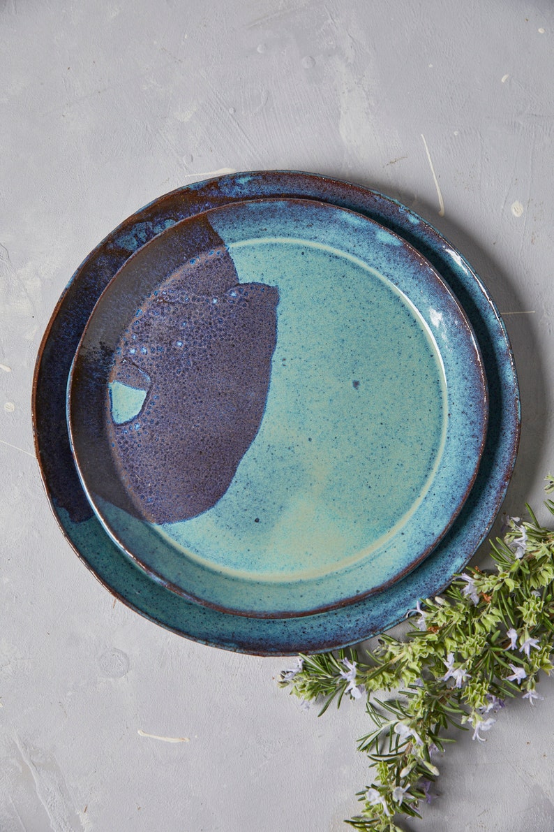 May include: Two ceramic plates, one nested inside the other. The top plate is a light blue with a dark blue and brown glaze. The bottom plate is a dark blue with a brown glaze.