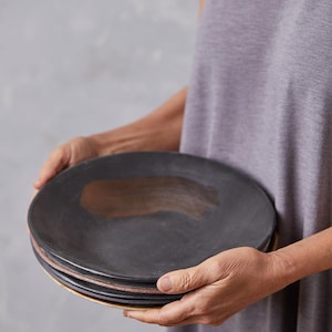 May include: A stack of four black ceramic plates with a brown glaze on the bottom. The plates are being held by a person's hands.