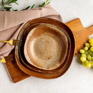 Op de afbeelding: Een set van drie keramische borden met een gespikkelde, aardse afwerking. Twee gouden vorken liggen op een roze linnen servet. Een houten snijplank en een tros groene druiven maken het stilleven compleet.