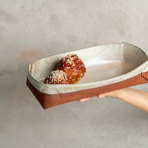May include: A rectangular ceramic serving dish with a red-brown exterior and a cream-coloured interior. The dish holds two pastries topped with nuts. The dish is held by a person's hand against a neutral background.