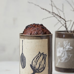 Op de afbeelding: Een keramische container met een gebakken product erin, met een zwart bloemenontwerp. De container is beige met bruine randen. Een andere container met een wit bladerontwerp staat op de achtergrond. Het gebakken product is donkerbruin.