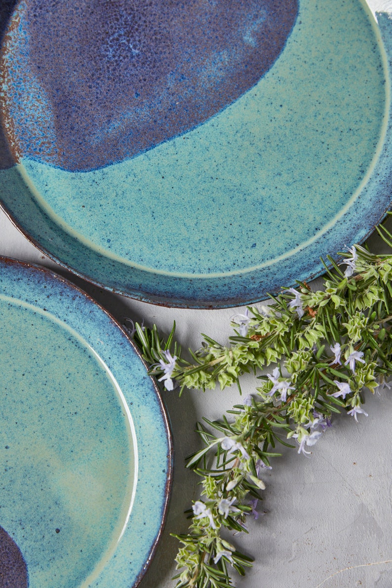 May include: Two blue and green ceramic plates with a speckled glaze. The plates are stacked on top of each other. A sprig of rosemary with purple flowers is in the background.
