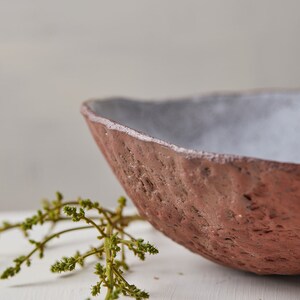 Op de afbeelding: Een rustieke, handgemaakte keramische kom met een ruw, getextureerd exterieur en een glad, wit interieur. De kom is gedeeltelijk gevuld met een takje groen gebladerte.