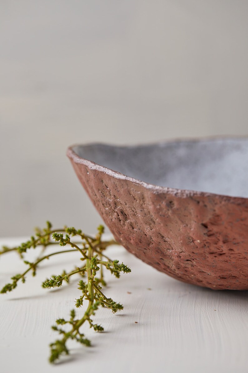 Op de afbeelding: Een rustieke, handgemaakte keramische kom met een ruw, getextureerd exterieur en een glad, wit interieur. De kom is gedeeltelijk gevuld met een takje groen gebladerte.