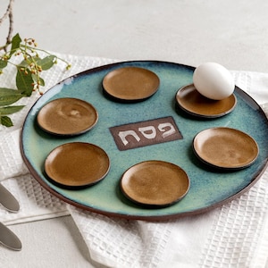 May include: A ceramic Seder plate with a teal glaze and brown accents. The plate holds six small brown dishes, an egg, and a central plaque with Hebrew text. Two forks and greenery are on the side.
