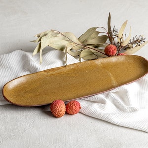 May include: A long, oval-shaped ceramic serving platter in a warm, golden-brown hue. The platter rests on a white waffle-weave cloth, with dried foliage and two red lychees. The platter's dimensions are approximately 16 inches long.