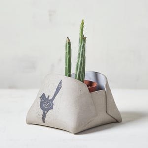 May include: A light beige ceramic planter with a folded, angular shape and a dark gray graphic print of a bird perched on a branch. The planter holds two green, slender, upright cacti in a small brown pot, visible within the open top section.