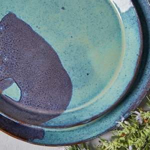 May include: Two ceramic plates, one stacked on top of the other. The top plate is a light blue with a dark blue and brown splatter pattern. The bottom plate is a darker blue with a brown splatter pattern. The plates are on a white surface with green rosemary sprigs.